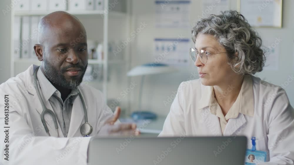 African American male doctor discussing medical data on laptop screen with middle-aged Caucasian female colleague while working together in clinic African American male doctor discussing medical data on laptop screen with middle-aged Caucasian female colleague while working together in clinic