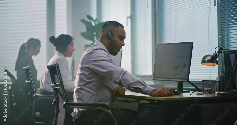 Multiethnic team of technical support operators working in modern call center office. African American helpdesk specialist talks on headset, uses computer, looks at camera providing customer service. Multiethnic team of technical support operators working in modern call center office. African American helpdesk specialist talks on headset, uses computer, looks at camera providing customer service.