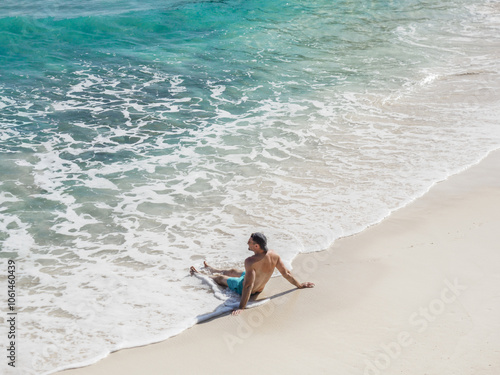 Handsome man sitting on stunning white sand beach with palm trees and rocks in the background. Beautiful views and sights of Seychelles. Clear, warm sunlight. Outdoor. Concept of leisure and travel