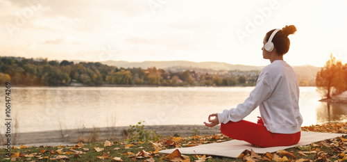 Bild auf Leinwand Female person in headphones sits in lotus pose in front of autumn river in nature and meditating during sunset