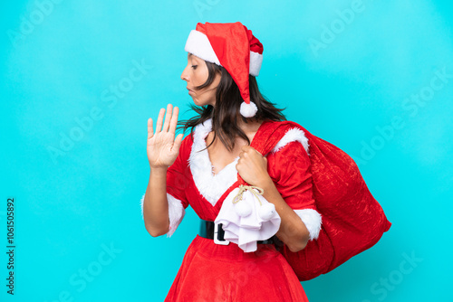 Young hispanic woman dressed as mama noel holding a sack isolated on blue background making stop gesture and disappointed