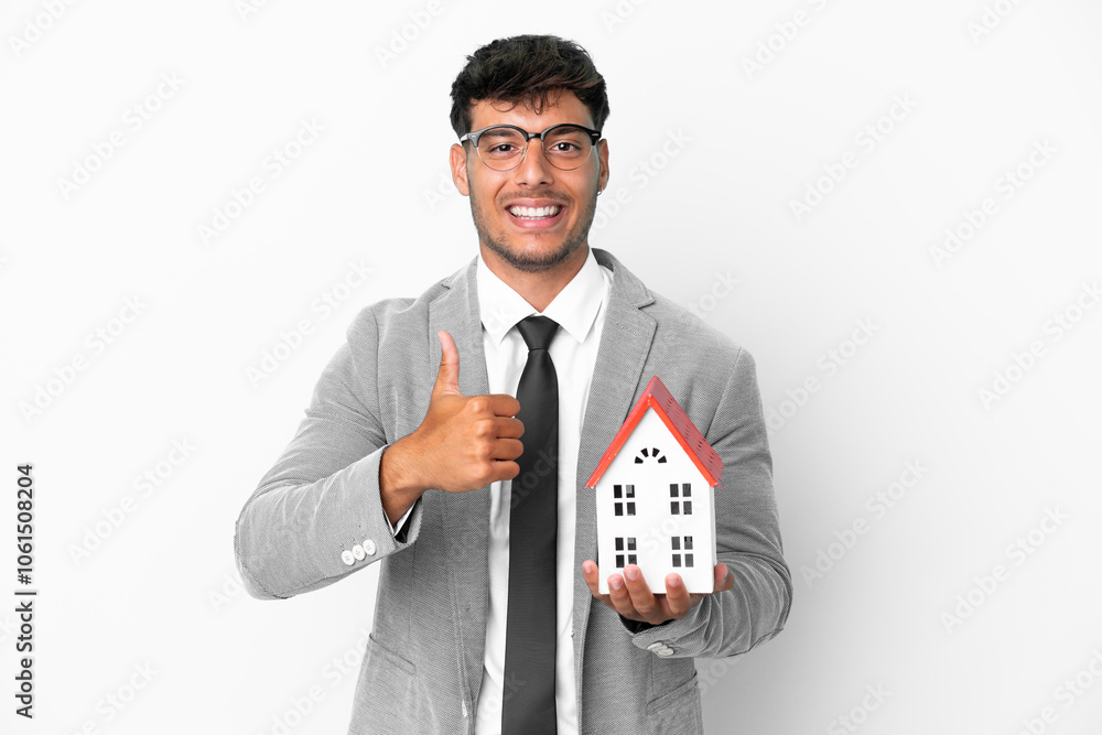 Business man holding a house isolated on blue background giving a thumbs up gesture Business man holding a house isolated on blue background giving a thumbs up gesture
