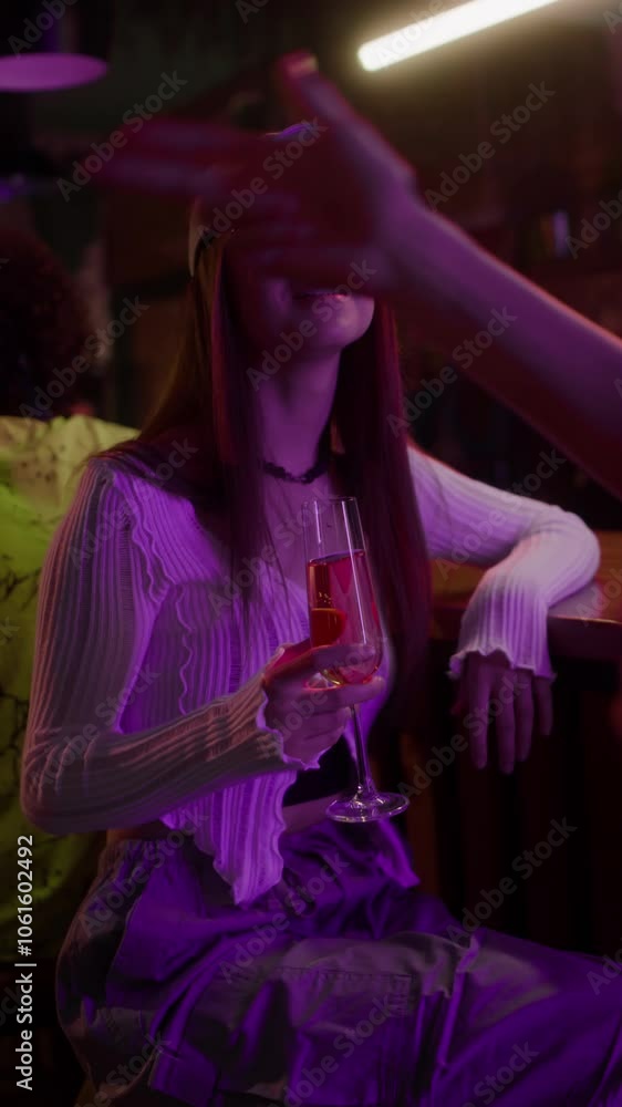 Vertical shot of long-haired girl in cap with glasses holding glass of champagne talking to unknown man while sitting at bar counter Vertical shot of long-haired girl in cap with glasses holding glass of champagne talking to unknown man while sitting at bar counter