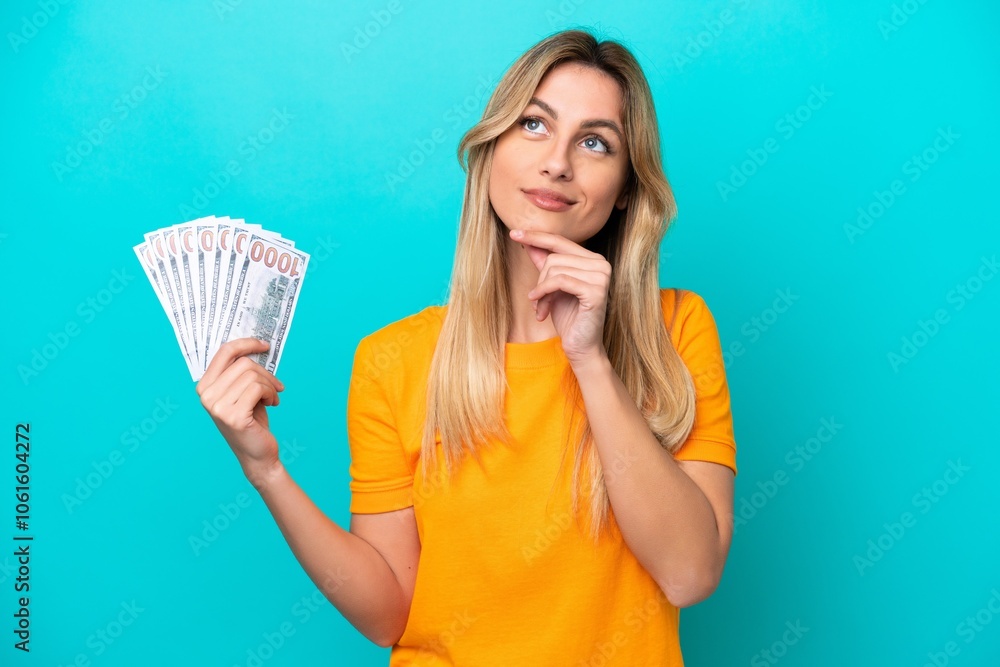 Young Uruguayan woman taking a lot of money isolated on blue background having doubts while looking up Young Uruguayan woman taking a lot of money isolated on blue background having doubts while looking up