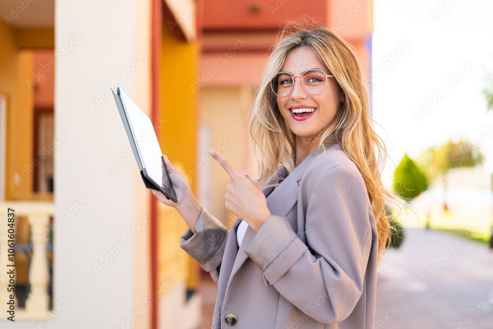 Pretty blonde Uruguayan woman holding a tablet at outdoors and pointing it Pretty blonde Uruguayan woman holding a tablet at outdoors and pointing it