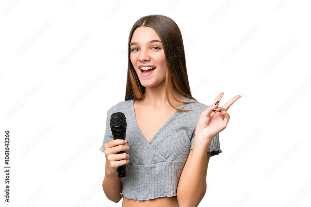 Teenager singer girl picking up a microphone over isolated background smiling and showing victory sign Teenager singer girl picking up a microphone over isolated background smiling and showing victory sign