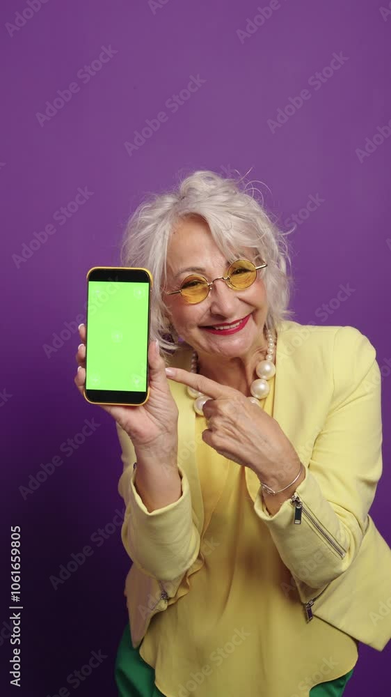 Stylish senior woman presenting smartphone with green screen on purple background Stylish senior woman presenting smartphone with green screen on purple background