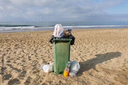 An overflowing green garbage bin on a sandy beach with scattered trash bags around it, set against a cloudy oceanfront background, highlighting beach maintenance and environmental concerns
