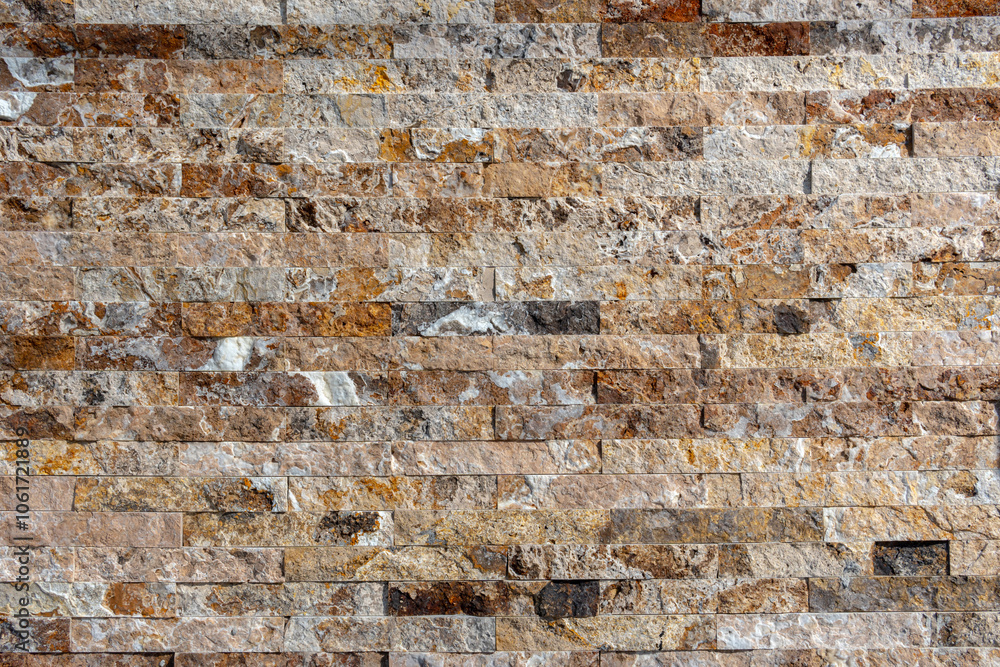 Fototapeta premium Wall made of stacked stone tiles in various shades of brown, beige, gray, and white. The stones are arranged horizontally with a rough texture, giving the wall a rustic and natural appearance.