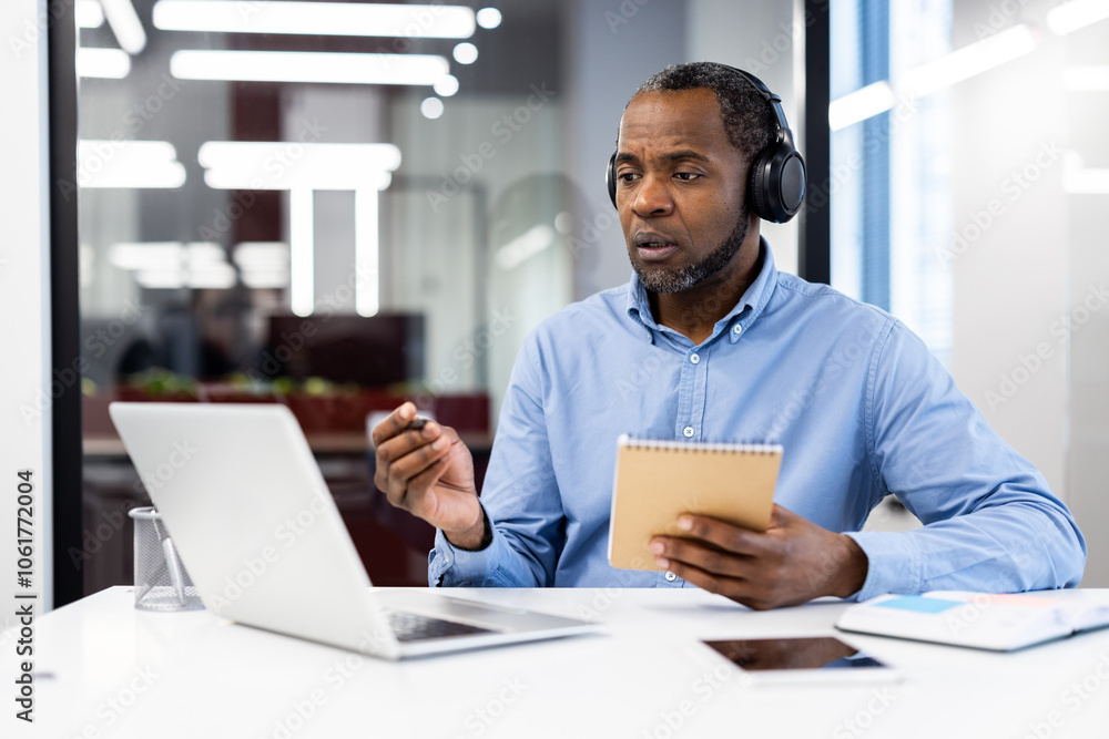 Mature african american businessman in video conference, wearing headphones, holding notepad while talking to laptop. Engaged in virtual meeting in modern office. Represents remote work, technology Mature african american businessman in video conference, wearing headphones, holding notepad while talking to laptop. Engaged in virtual meeting in modern office. Represents remote work, technology
