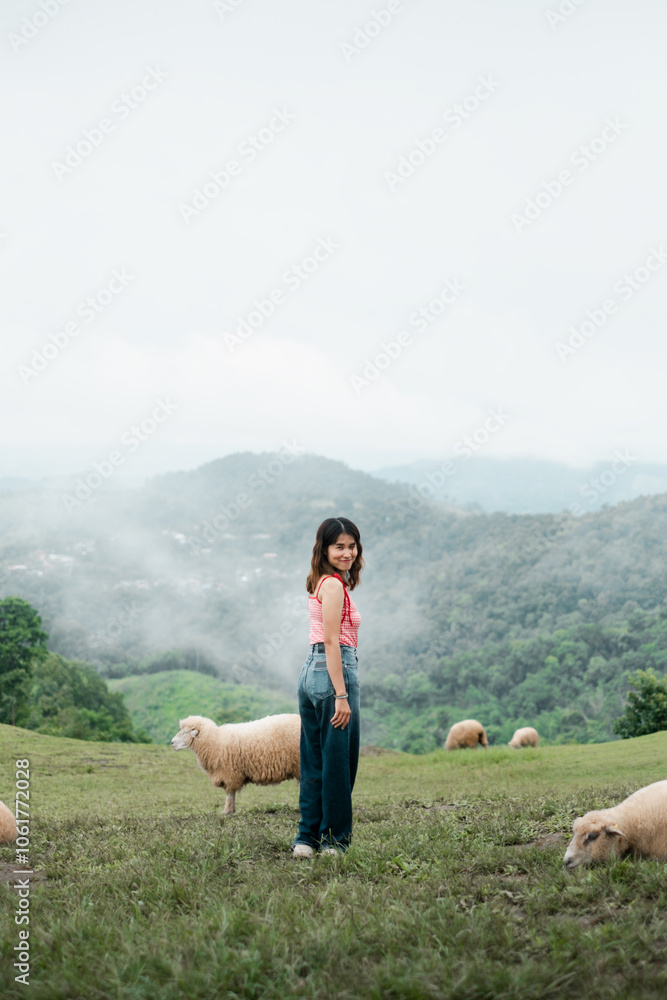 A woman stands in a green pasture with sheep, surrounded by misty mountains, creating a serene and pastoral scene.