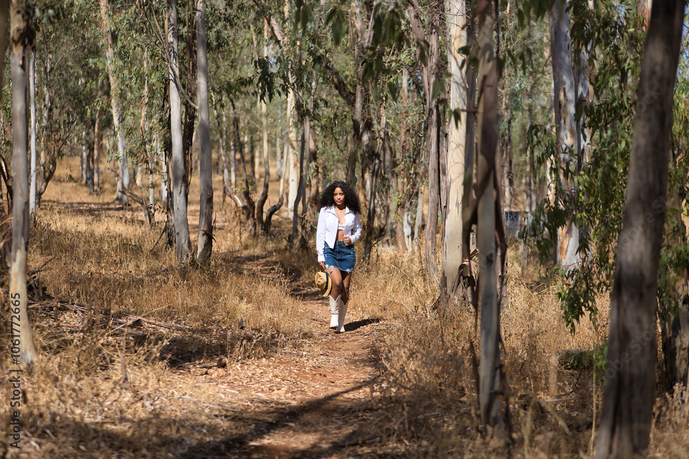 Obraz premium Latin woman, young, beautiful, dark-haired, with curly hair, with a white shirt and top, denim skirt and hat, walking alone and calmly through a eucalyptus forest. Concept peace, relaxation.