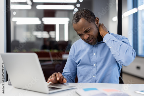 Mature african american businessman experiencing neck pain at desk using laptop in office. Display of discomfort, stress in work environment.