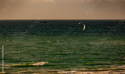 Watersport: kitesurfer riding the ocean waves with a red kite in strong wind conditions, creating splashes as they speed across the water