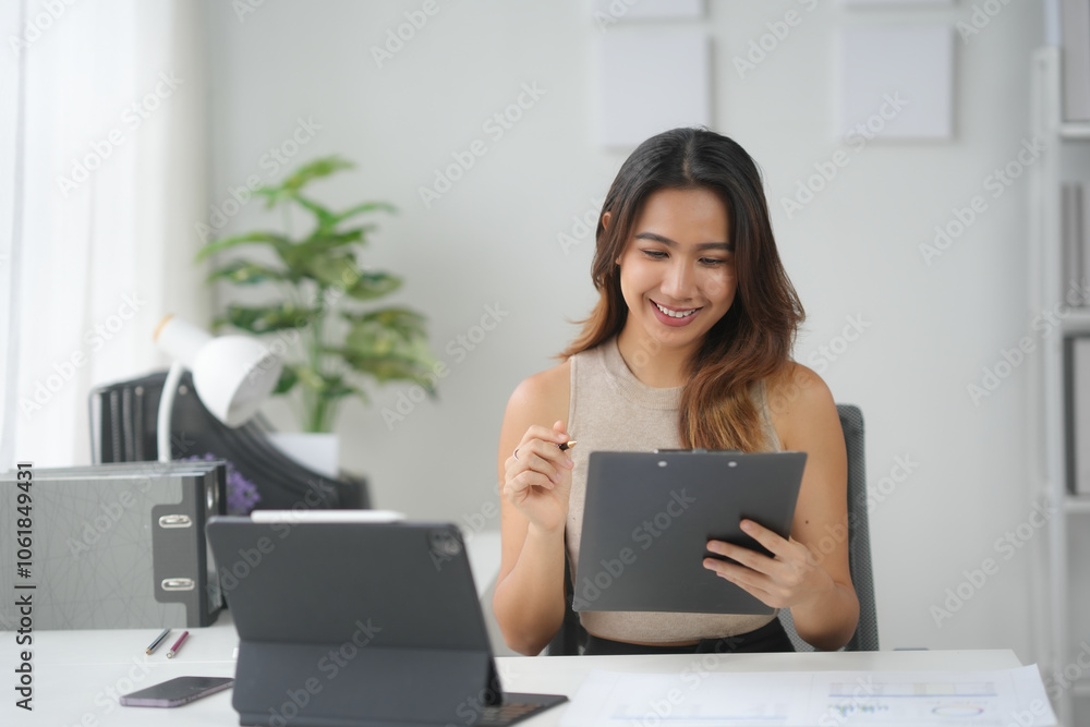 Businesswoman working at desk with tablet and clipboard in a bright office space. She is smiling and focused on her tasks. Businesswoman working at desk with tablet and clipboard in a bright office space. She is smiling and focused on her tasks.