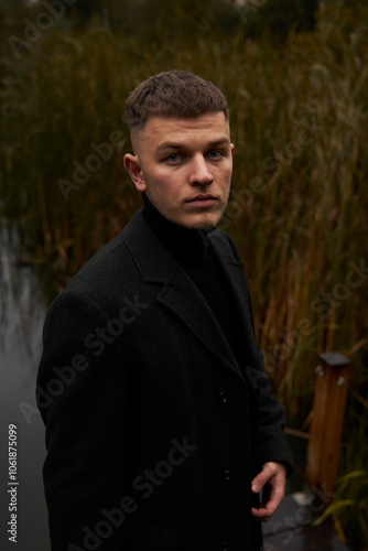 A European man in a black coat walks in the park near the water at dusk in autumn