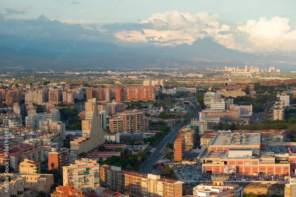 A panoramic aerial view of a bustling city with modern buildings, a busy road, and a scenic mountain range in the background under a partly cloudy sky. Alicante, Spain A panoramic aerial view of a bustling city with modern buildings, a busy road, and a scenic mountain range in the background under a partly cloudy sky. Alicante, Spain