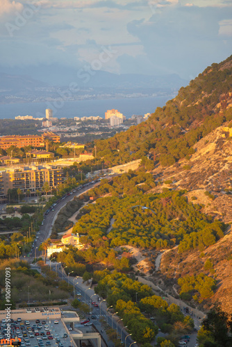 A picturesque urban landscape featuring a winding road, lush greenery, and a coastal cityscape with mountains in the background, captured during golden hour.