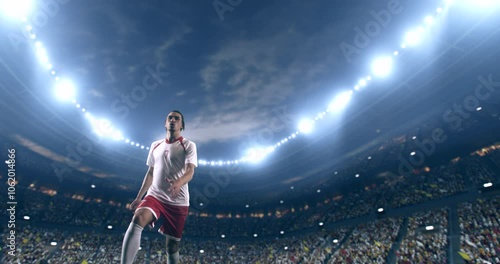 Football player, wearing red and white uniform, kicks the ball in a jump, in slow motion on the professional football stadium with animated crowd.