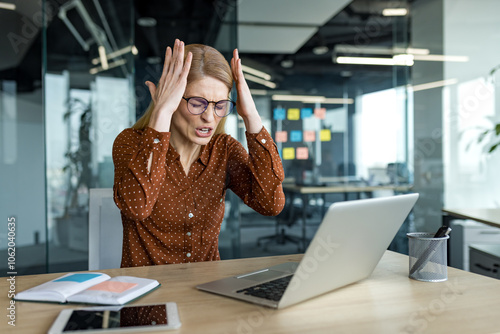 Frustrated businesswoman sitting in modern office facing laptop, expressing stress. Desk features phone, notebook, suggesting overwhelming work pressure and deadline anxiety.