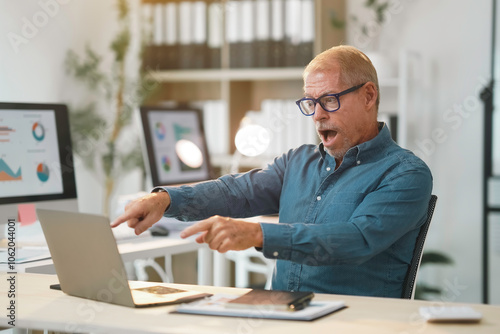 Surprised mature businessman pointing at laptop screen with a shocked expression, reacting to unexpected news or data