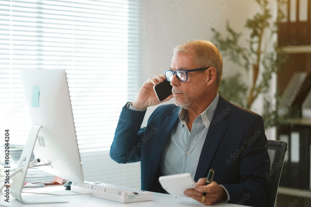 Focused mature businessman using smartphone and taking notes while working on computer in modern office Focused mature businessman using smartphone and taking notes while working on computer in modern office