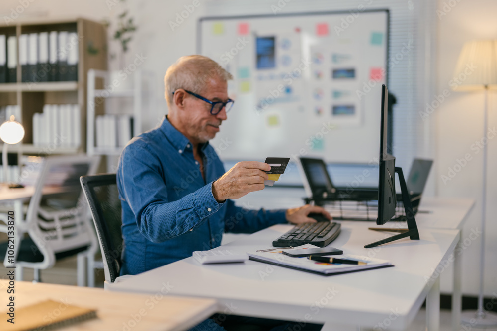 Senior businessman sitting at his desk in a modern office, holding a credit card and typing on his computer keyboard, making an online payment or purchase Senior businessman sitting at his desk in a modern office, holding a credit card and typing on his computer keyboard, making an online payment or purchase