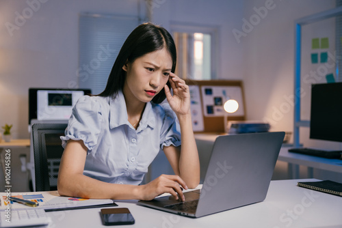 Young asian businesswoman is massaging her head because of a headache while working on a laptop sitting at her desk in a modern office at night