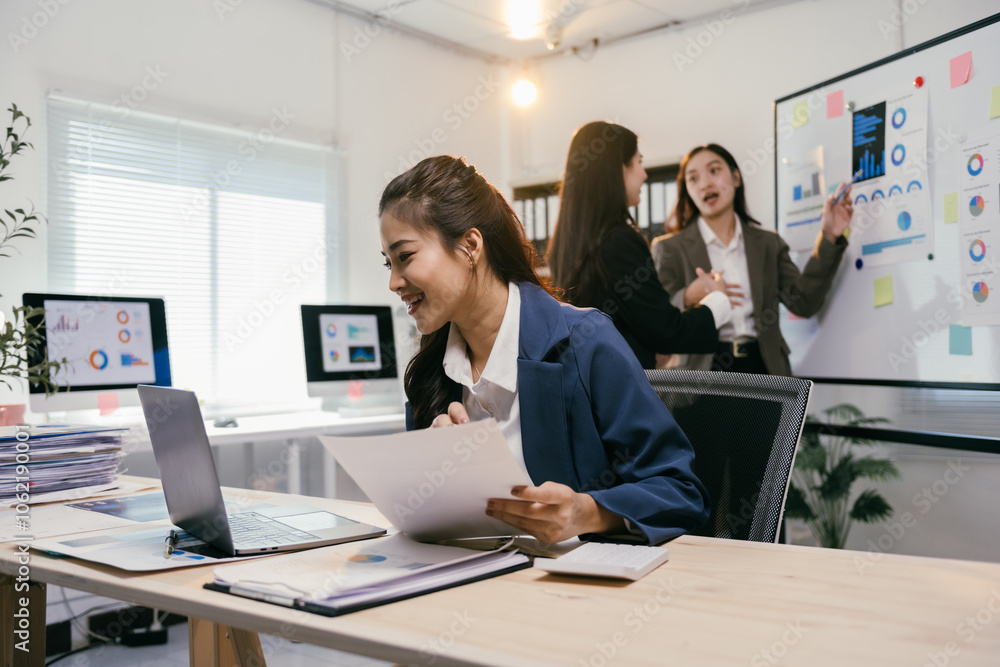 Businesswomen are collaborating on a project, analyzing financial data on a laptop and documents, while other colleagues discuss strategy near a whiteboard with charts and graphs Businesswomen are collaborating on a project, analyzing financial data on a laptop and documents, while other colleagues discuss strategy near a whiteboard with charts and graphs