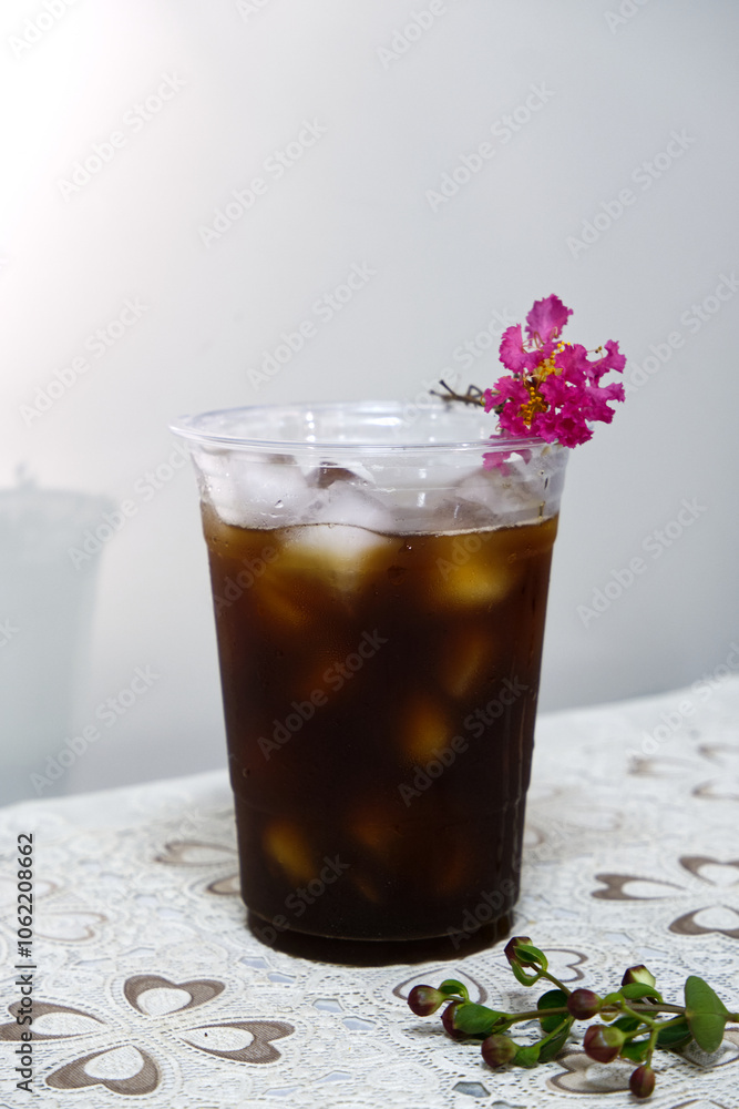 Iced coffee glass with pink flower placed on top with natural light. Front view iced coffee plastic cup for summer. Concept of takeaway, cold drink packaging, coffee, energy, and relaxing at a cafe. Iced coffee glass with pink flower placed on top with natural light. Front view iced coffee plastic cup for summer. Concept of takeaway, cold drink packaging, coffee, energy, and relaxing at a cafe.
