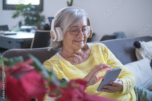 Smiling relaxed senior woman wearing headphones sitting on sofa at home using smart phone looking at social news