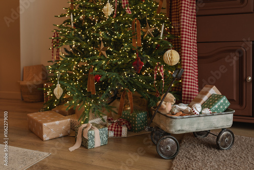 Wrapped Christmas gifts under an illuminated Christmas tree and in a toy wagon in a living room photo