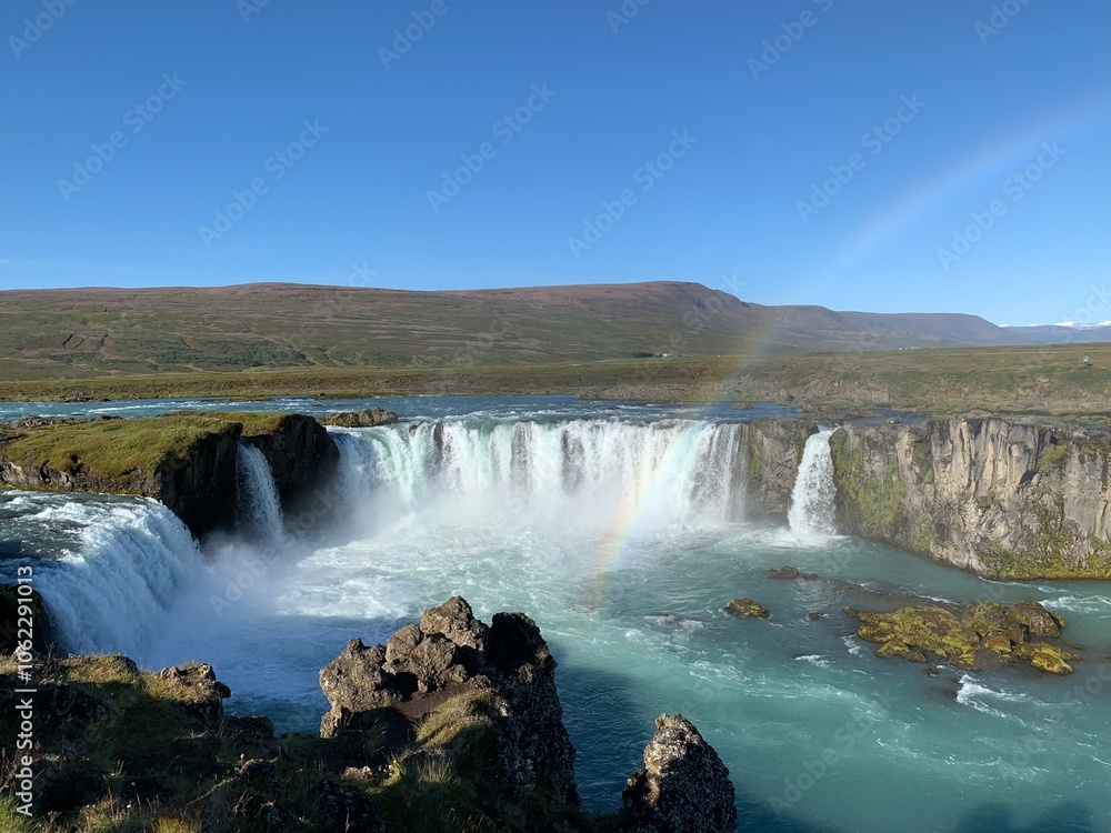 Fototapeta premium Goðafoss waterfall in Iceland, known as the 'Waterfall of the Gods,' cascades powerfully into a deep river. The stunning waterfall is framed by rugged rocks and surrounded by vast, serene landscapes.