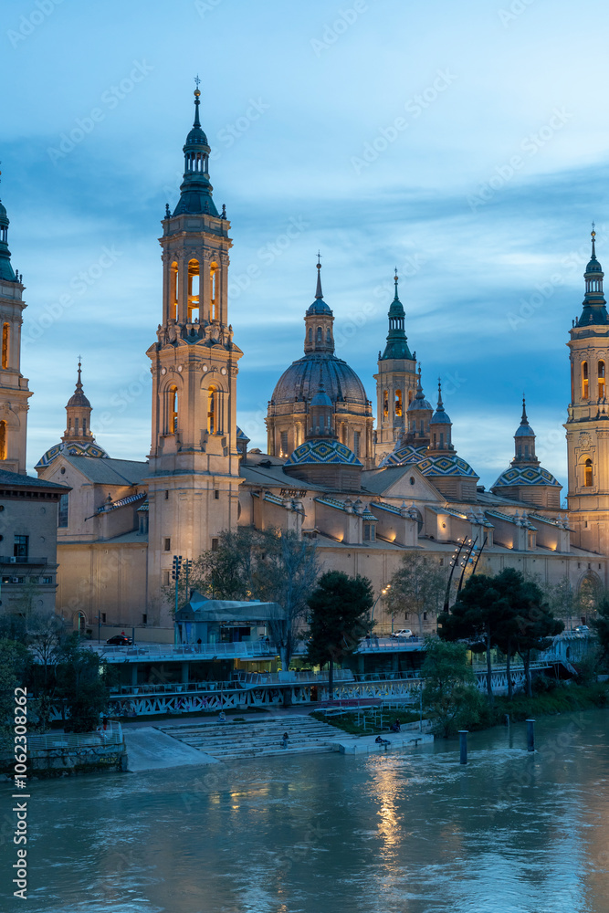 Illuminated Basilica del Pilar in Zaragoza, Spain, at dusk, with serene reflections on the Ebro River. Ideal for travel, architecture, and cultural themes. Illuminated Basilica del Pilar in Zaragoza, Spain, at dusk, with serene reflections on the Ebro River. Ideal for travel, architecture, and cultural themes.