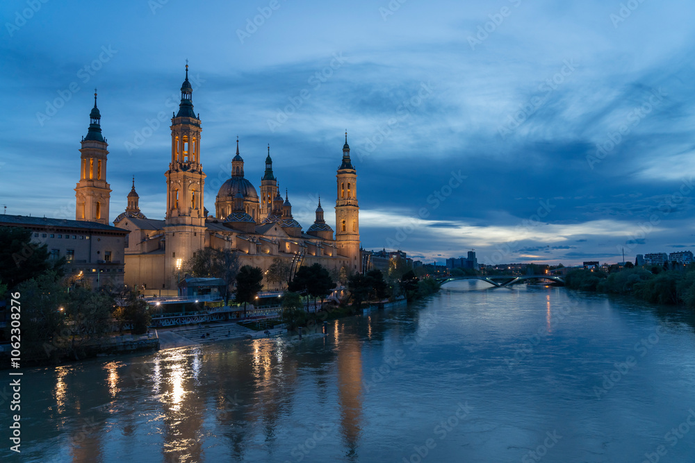 Illuminated Basilica del Pilar in Zaragoza, Spain, at dusk, with serene reflections on the Ebro River. Ideal for travel, architecture, and cultural themes. Illuminated Basilica del Pilar in Zaragoza, Spain, at dusk, with serene reflections on the Ebro River. Ideal for travel, architecture, and cultural themes.