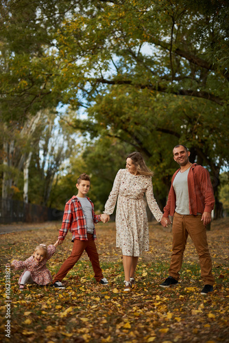 Happy family walking on green grass strewn with fallen leaves in Park in autumn. Parents, two children are walking in park at sunset with holding hands. Family, child, walk through Autumn Park.