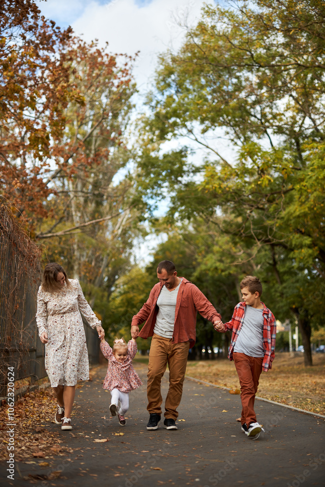 Fototapeta premium Happy family walking on green grass strewn with fallen leaves in Park in autumn. Parents, two children are walking in park at sunset with holding hands. Family, child, walk through Autumn Park.