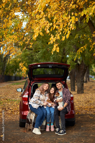 Happy parents with daughter and cute dog Jack Russel terrier sitting in car trunk on autumn day. Long auto journey break.