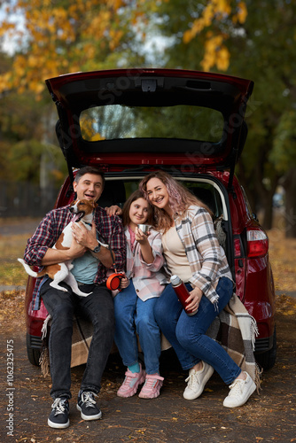 Happy parents with daughter and cute dog Jack Russel terrier sitting in car trunk on autumn day. Long auto journey break.