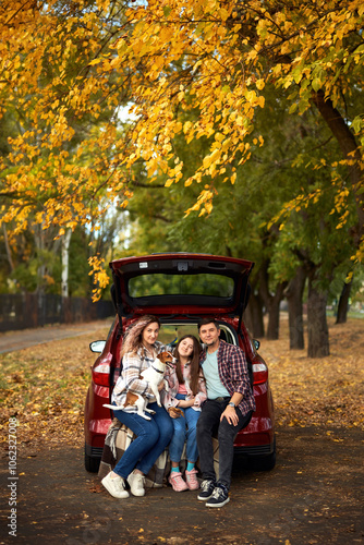 Happy parents with daughter and cute dog Jack Russel terrier sitting in car trunk on autumn day. Long auto journey break.