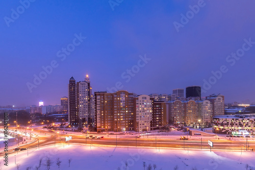 The city skyline features tall buildings glowing with lights at dusk. Snow blankets the ground and roads, creating a wintry atmosphere around the bustling urban landscape.. photo