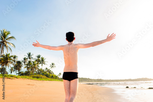 Young man wearing  a swimwear  is walking on the beach sand with his arms wide open, expressing well-being and gratitude during his beach trip.  photo