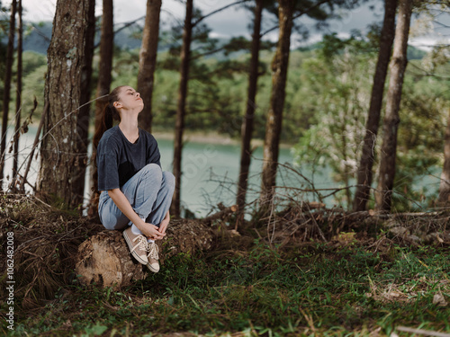 Thoughtful young woman sitting on a log in the forest, enjoying serene moments and natural beauty, surrounded by lush greenery and calm water