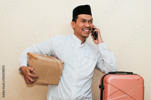 Smiling Southeast Asian muslim man carrying cardboard box and suitcase while holding mobile phone taking a call. Isolated on white background. Ideal for concepts of Ramadan and mudik travel.