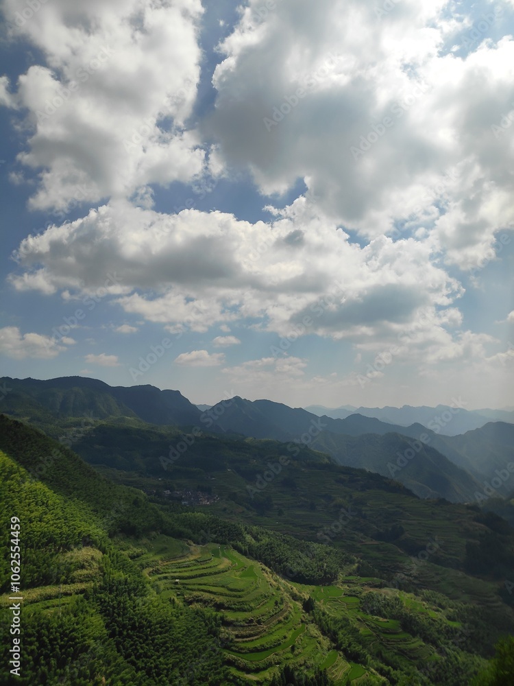 Fototapeta premium This breathtaking image captures a panoramic view of expansive mountain ranges with lush, terraced fields below under a beautiful sky dotted with fluffy white clouds.