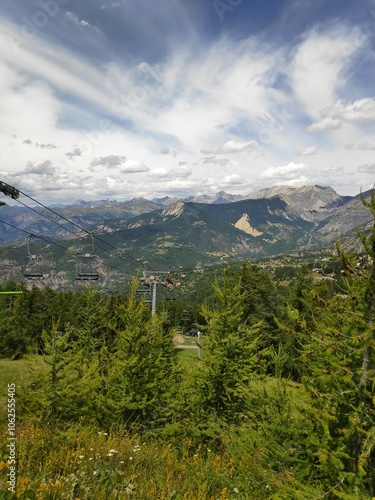 The image presents a dramatic view from mountain slopes, with a dynamic cloudy sky overhead and evergreen trees in the foreground, offering a sense of adventure.