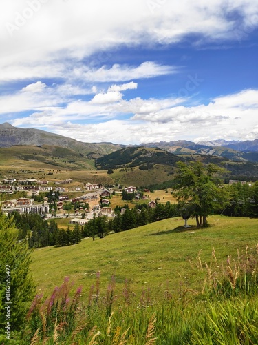 This image showcases a picturesque mountain village nestled amidst lush greenery, with a vast blue sky above and distant mountain ranges creating a serene scene.