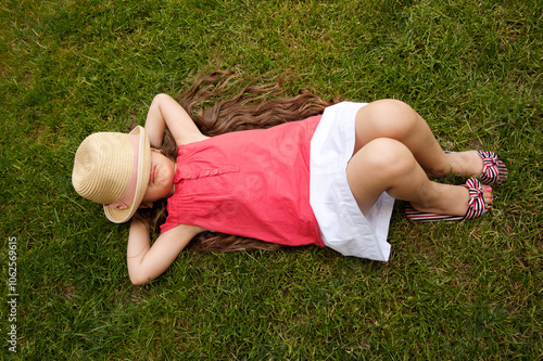 A young girl with long hair wearing a red top and white skirt lies relaxed on green grass, with a straw hat covering her face, capturing a serene summer moment in nature.