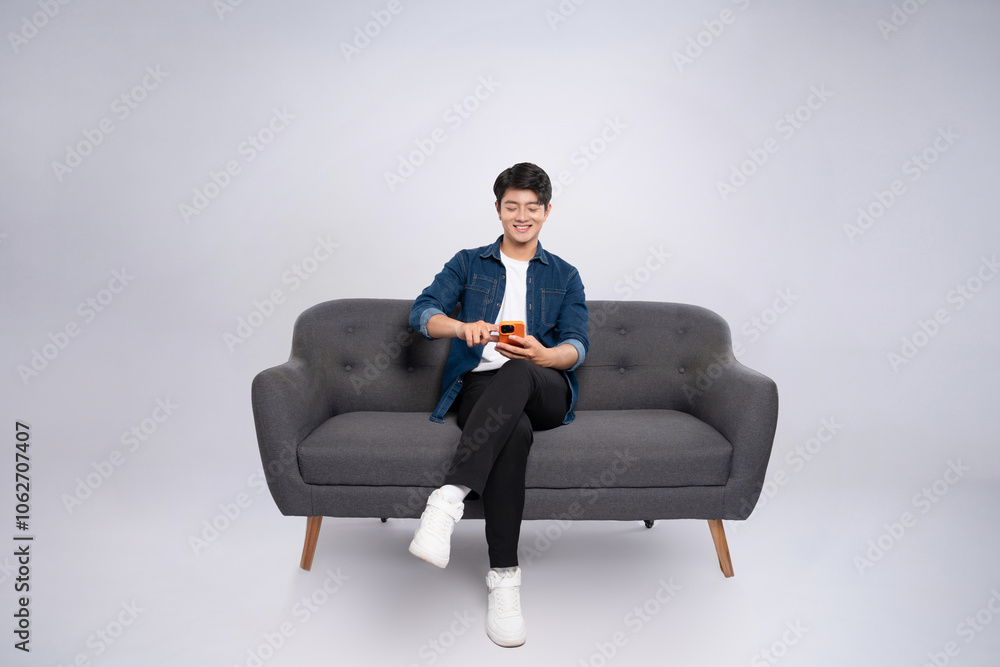 Full body image of young Asian man using phone sitting on sofa on white background Full body image of young Asian man using phone sitting on sofa on white background