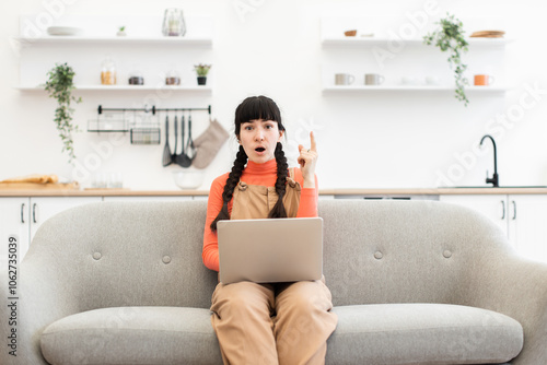 Caucasian woman in casual attire working on laptop from home. Young adult with braided hairstyle shows thoughtful expression while seated on cozy sofa in modern living room.